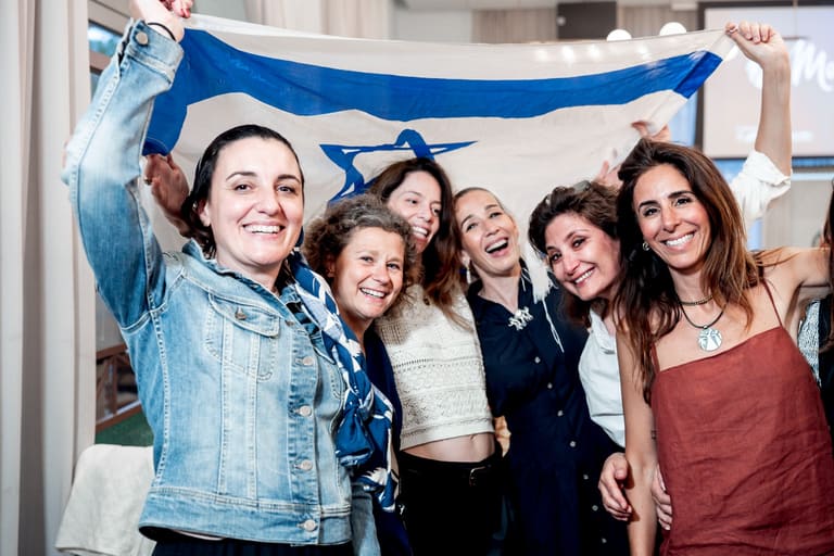 Six mothers laughing together beneath a raised Israeli flag
