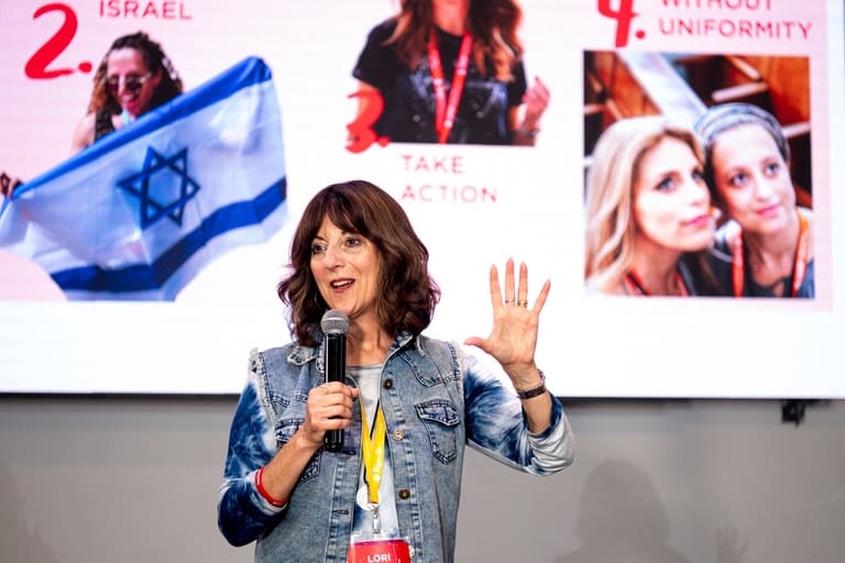 A Momentum educator teaching on stage with a microphone, gesturing in front of a screen of photos from Israel