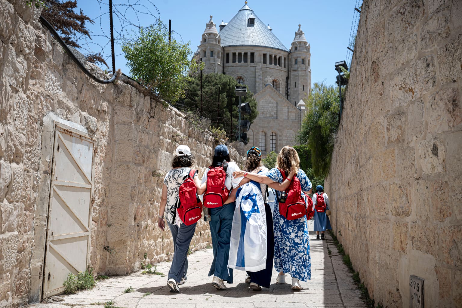 Four Jewish mothers with red Momentum backpacks, arms around each other and draped in an Israeli flag, walking up a Jerusalem alley toward Dormition Abbey